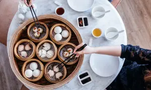Ręce sięgają po pyszne dim sum z bambusowego parownika. Na stole stoją miseczki z sosem i herbatą.