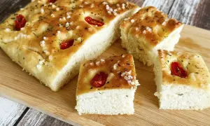 Oryginalna włoska focaccia przepis: puszyste ciasto z pomidorkami koktajlowymi, ziołami i solą morską na drewnianej desce.