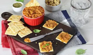 Pyszna pasta z suszonych pomidorów na krakersach, idealna na przekąskę. Obok miseczka z orzechami i szklanka wody.