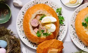 Pyszny żurek w chlebie z kiełbasą i jajkiem, udekorowany natką pietruszki. Obok wianek z pisankami i zajączek.