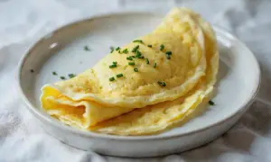 Delikatny omlet francuski, złożony na pół, posypany szczypiorkiem, na białym talerzu.
