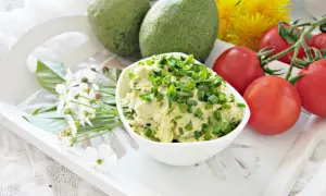 Pyszna pasta z awokado, jajkiem i szczypiorkiem, podana z pomidorkami i kwiatami na białej tacy.