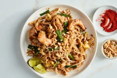 Pyszny sos pad thai z krewetkami, orzeszkami ziemnymi i limonką. Obok miseczka z chili i orzeszkami.