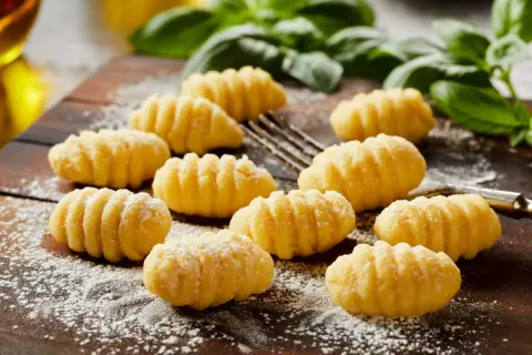 Pyszne, domowe **makaron gnocchi** ułożone na drewnianej desce, posypane mąką. W tle zielona bazylia i widelec.