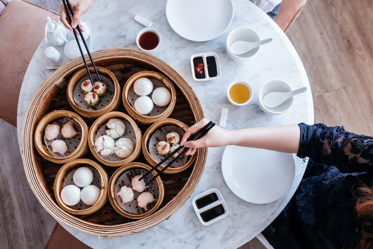 Ręce sięgają po pyszne dim sum z bambusowego parownika. Na stole stoją miseczki z sosem i herbatą.
