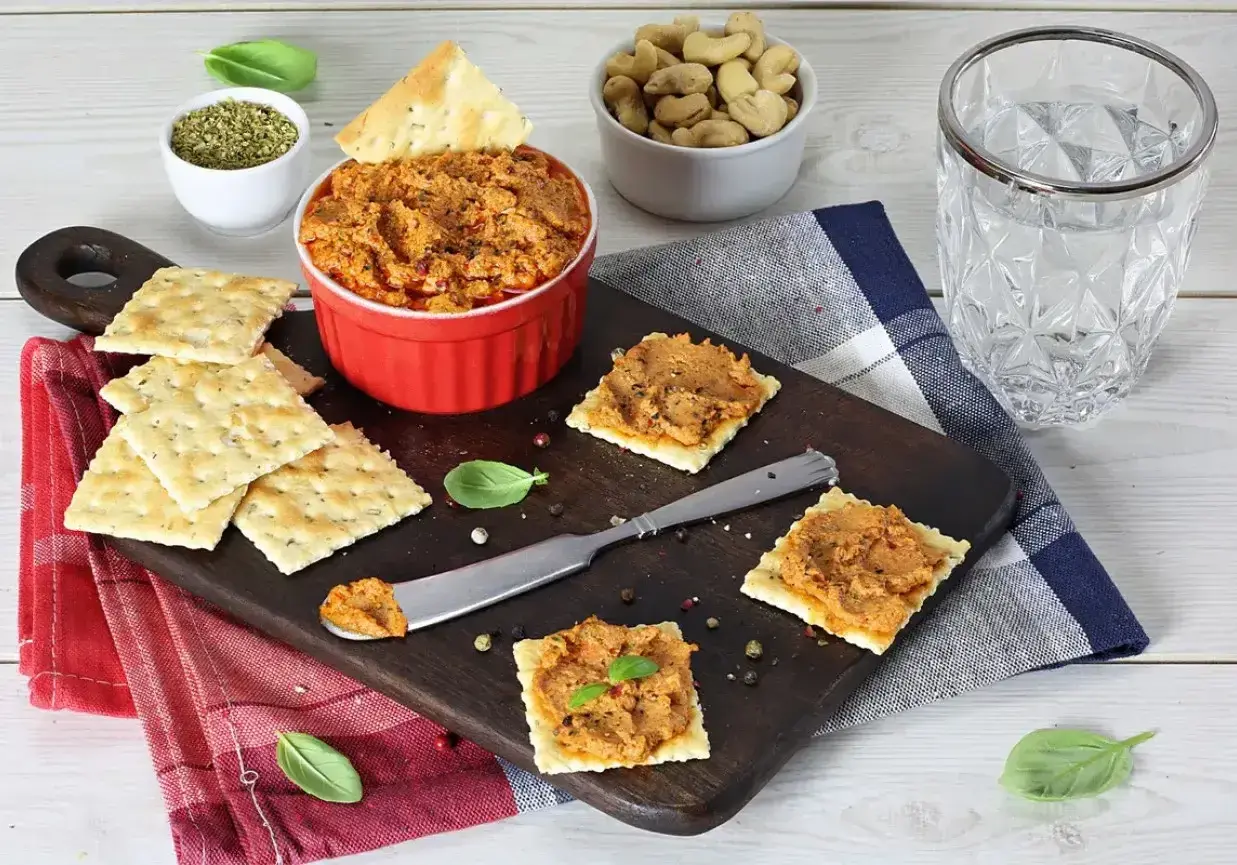 Pyszna pasta z suszonych pomidorów na krakersach, idealna na przekąskę. Obok miseczka z orzechami i szklanka wody.