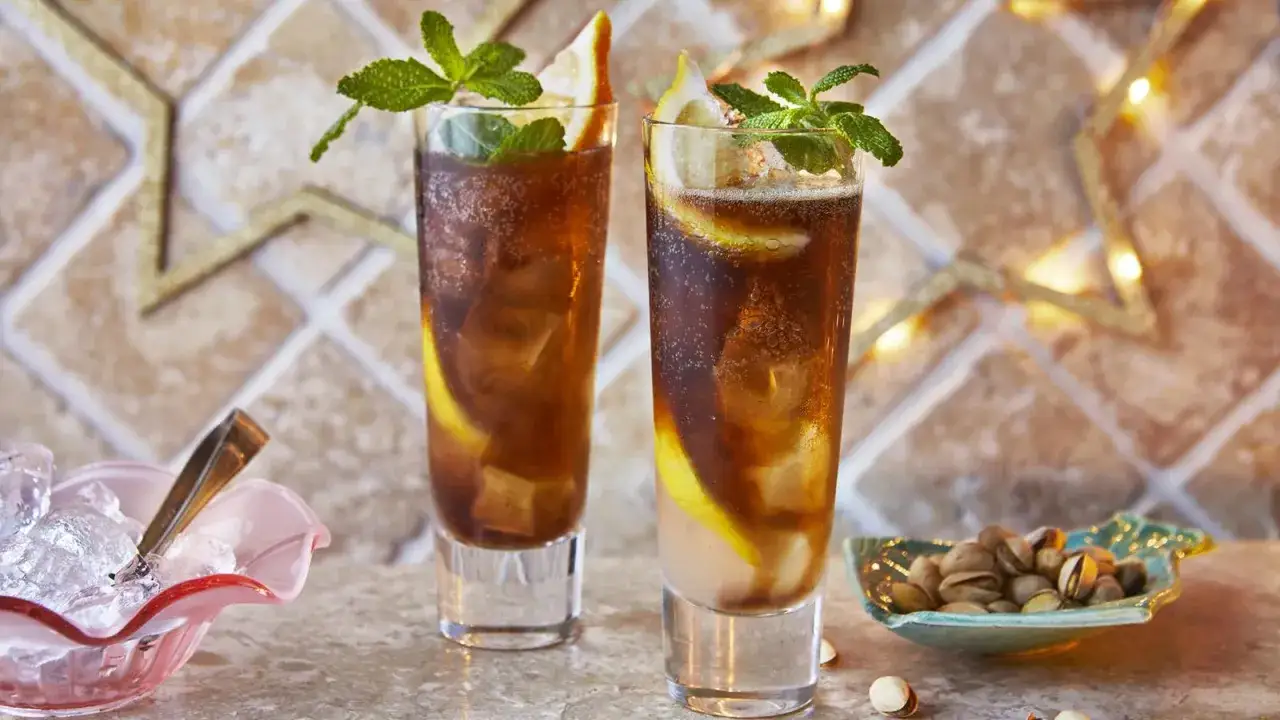 Dwa orzeźwiające drinki Long Island Ice Tea z cytryną i miętą, obok miseczki z pistacjami i kostkami lodu.