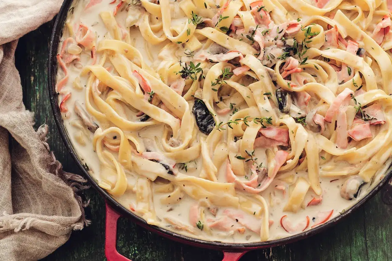 Pyszne danie z makaronem tagliatelle w kremowym sosie z kawałkami szynki i grzybów, udekorowane świeżym tymiankiem.