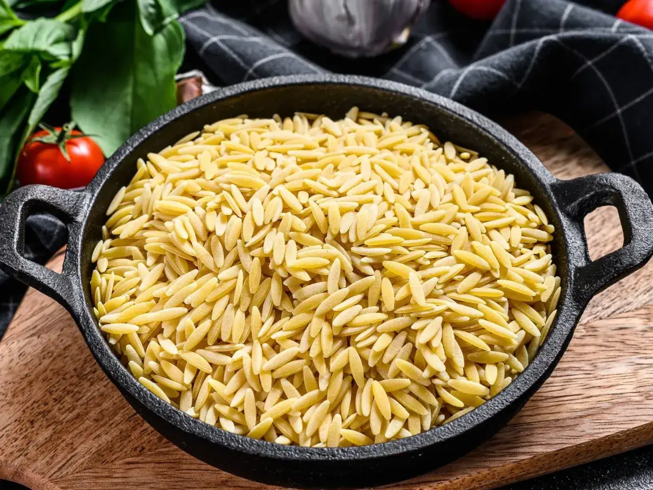 Makaron w kształcie ryżu, zwany orzo, w czarnej patelni. Obok pomidory i zielenina.