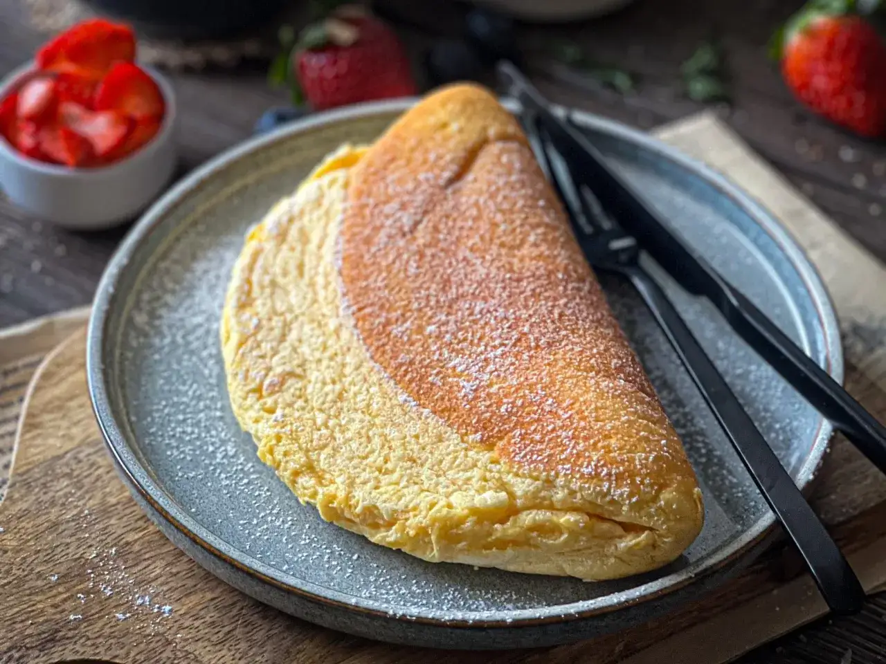 Puszysty omlet, lekko przyprószony cukrem pudrem, czeka na talerzu, by zachwycić swoim delikatnym smakiem. Obok leżą czarne sztućce.