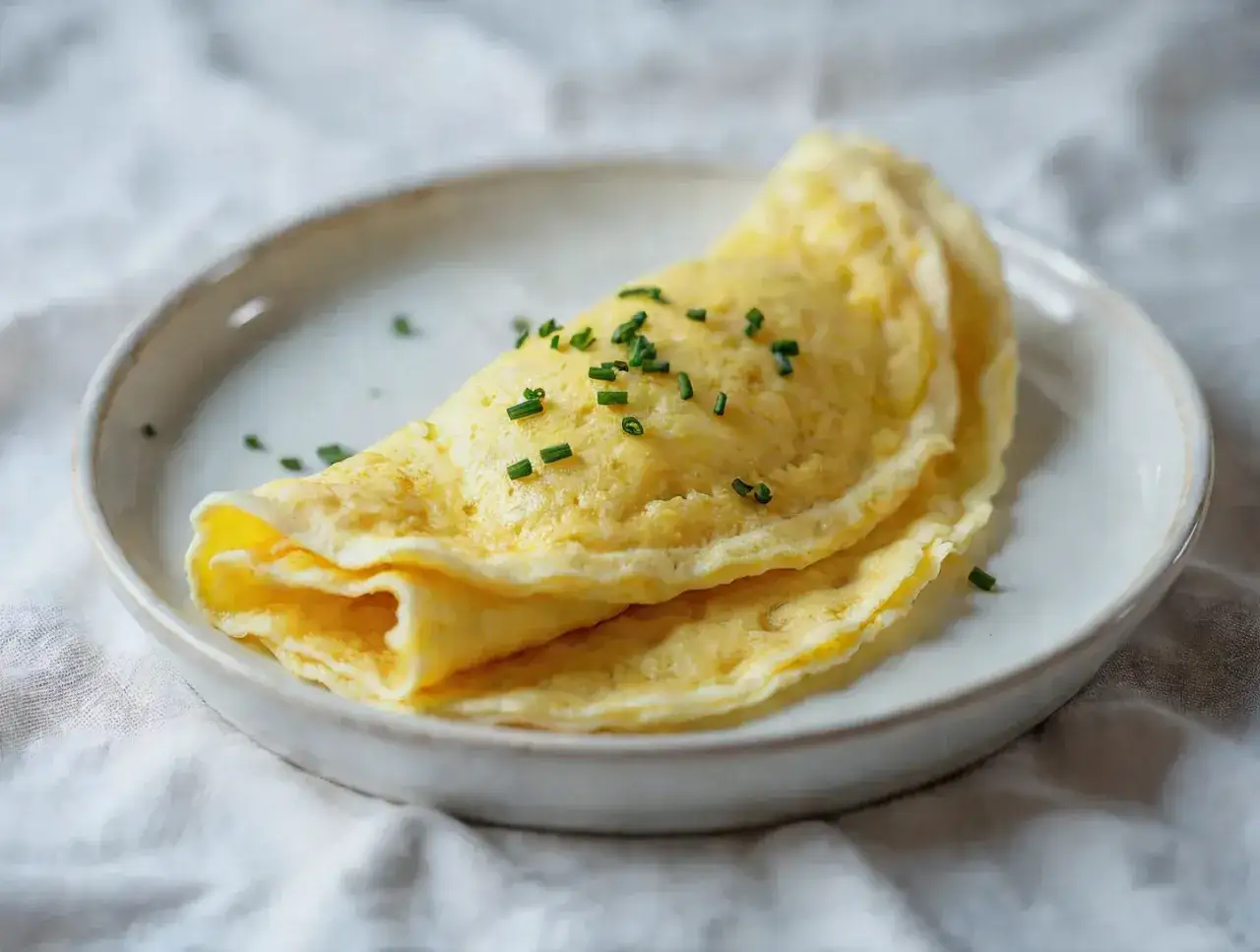 Delikatny omlet francuski, złożony na pół, posypany szczypiorkiem, na białym talerzu.