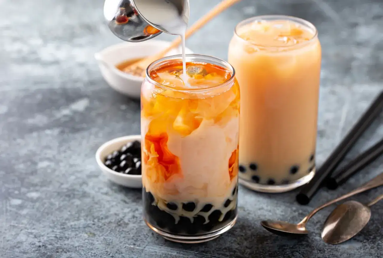 Jak zrobić bubble tea? Mleko wlewa się do szklanki z tapioką i pomarańczowym napojem. Obok stoi gotowy napój.