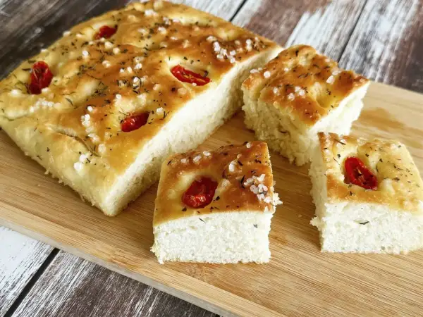 Oryginalna włoska focaccia przepis: puszyste ciasto z pomidorkami koktajlowymi, ziołami i solą morską na drewnianej desce.